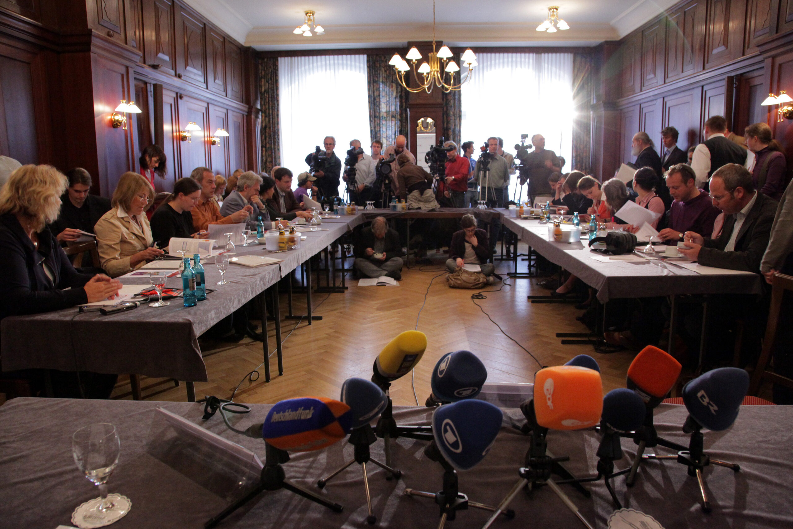 Bild von einer Pressekonferenz. Perspektive vom Podium aus, über eine Reihe von Mikrofonen hinweg, ins volle Publikum.