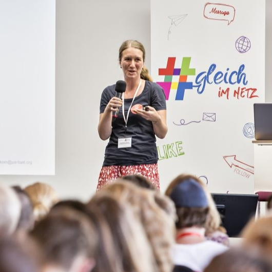 Frau mit Mikrofon steht vor Publikum. Im Hintegrund ein Rollup mit der Aufschrift #GleichImNetz